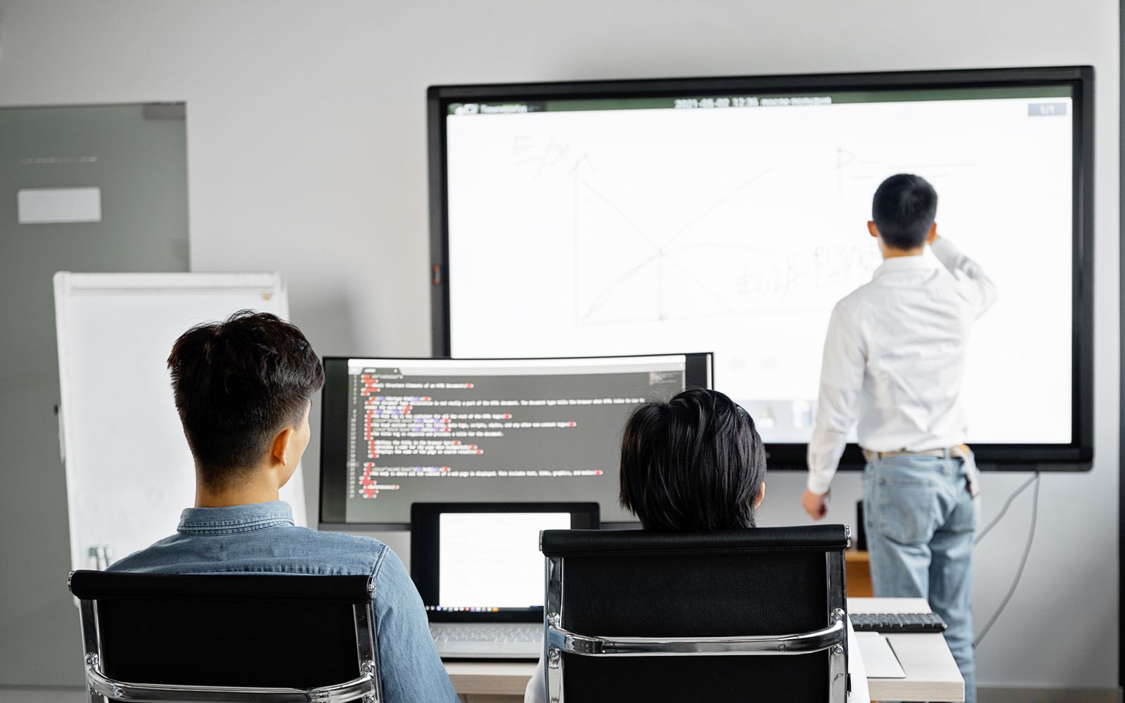 A presenter at a wall screen with two colleagues watching from their desks. Ask to see each test run live, not on a slide. Photo: Mikhail Nilov / Pexels.