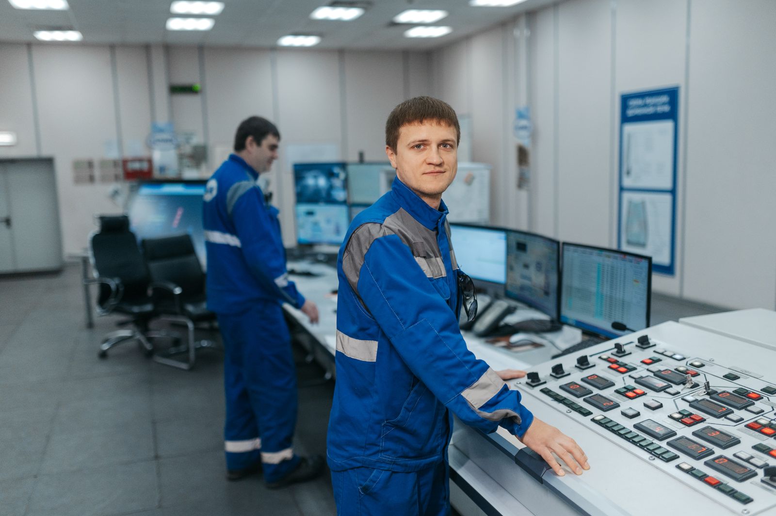 Two operators in blue overalls at a control room desk with monitors behind them. Photo: Sergey Sergeev / Pexels.