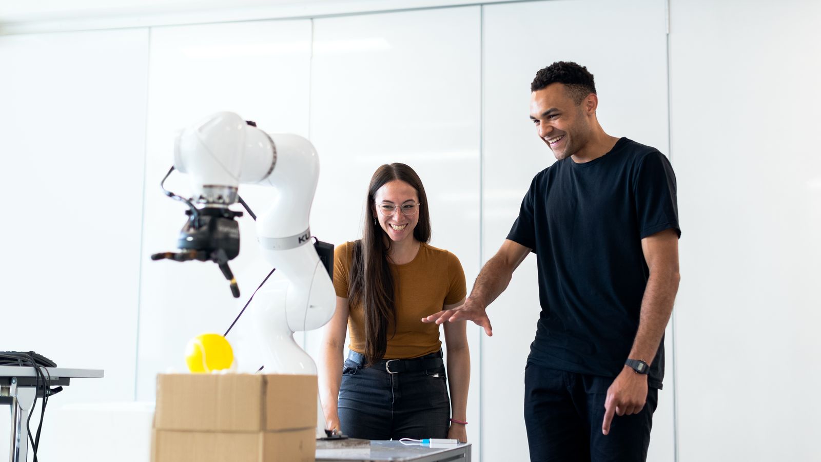 Two engineers watch a robot arm pick an object from a box. The machine does part of the work, and people still decide what it does. Photo: ThisIsEngineering / Pexels.