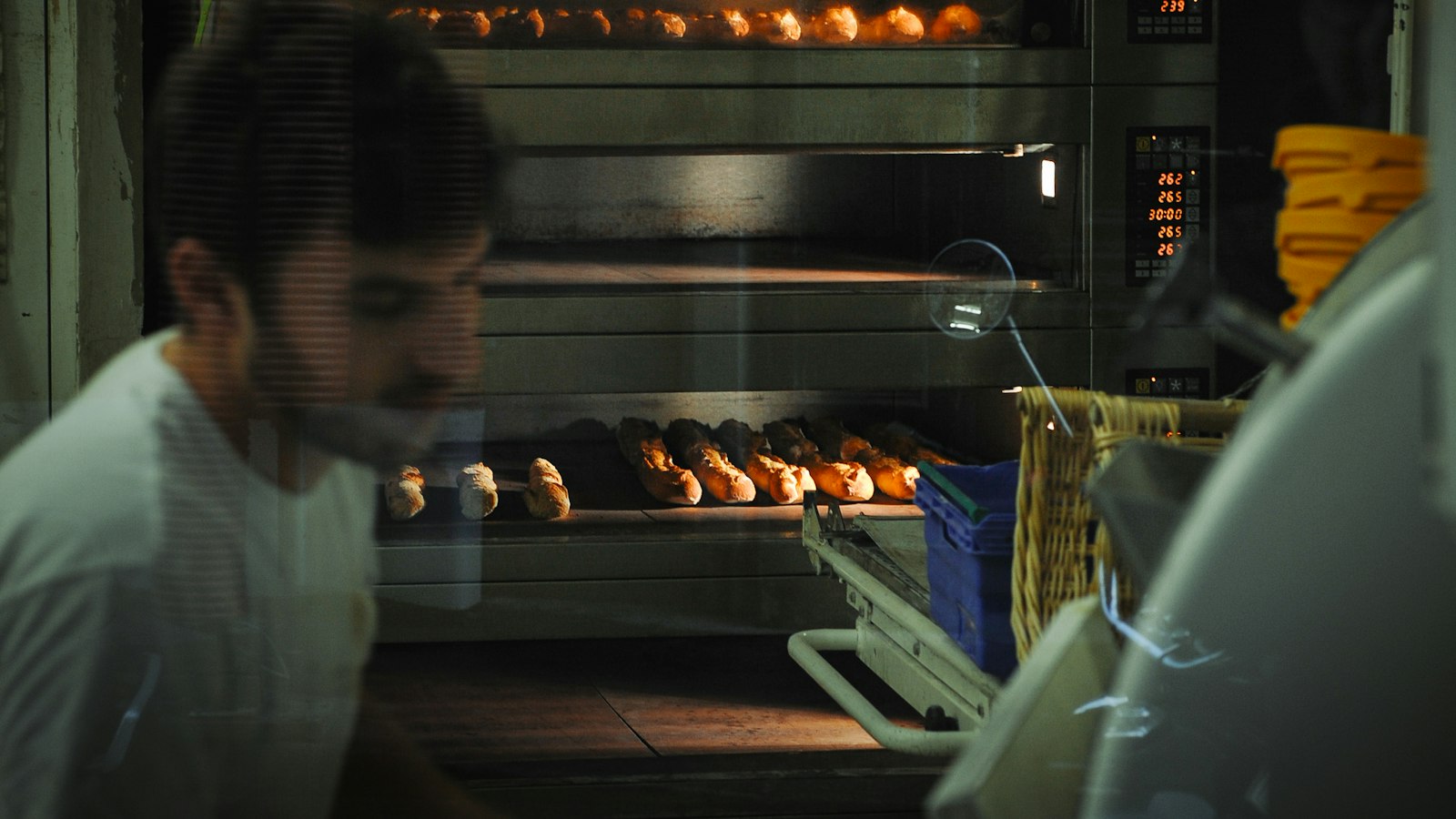 A baker at a deck oven in the dark, seen through the shop window, loaves on the hearth and the oven reading 262 degrees.