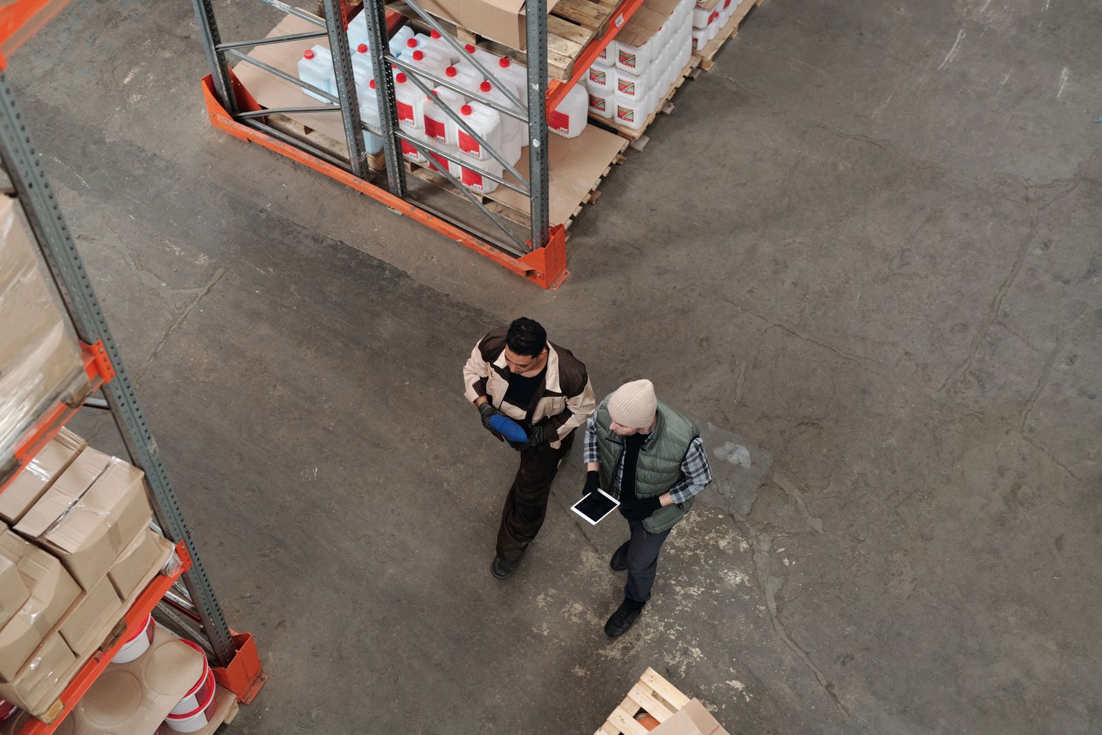 Two warehouse workers walk between pallet racks, seen from above; one checks a tablet, the other carries a scanner. Photo: Tiger Lily / Pexels.