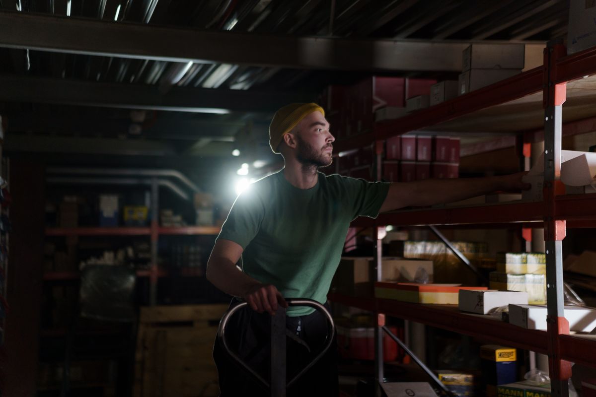 A warehouse worker in a green T-shirt and yellow beanie reaches for a box on a high shelf, one hand on a pallet truck, in a dim stockroom. Photo: cottonbro studio / Pexels.