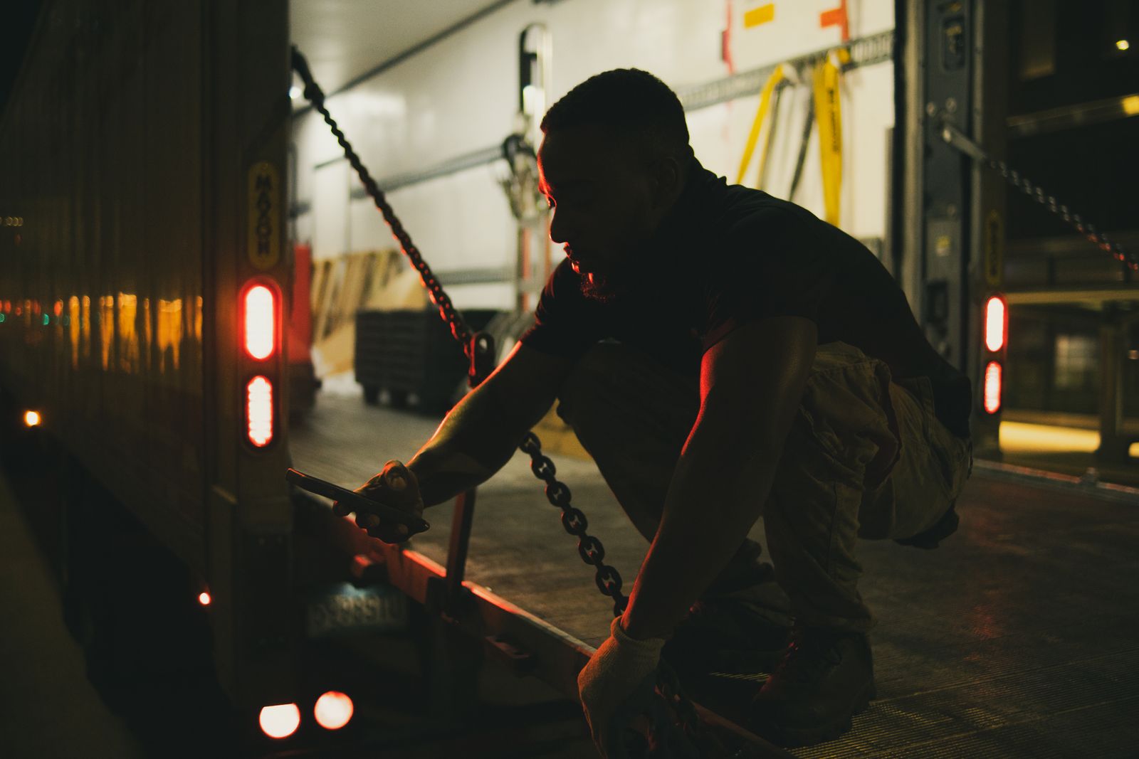 A worker crouches on the bed of a trailer at night, securing a load chain by the glow of the tail lights. Photo: Ian Findley / Pexels.