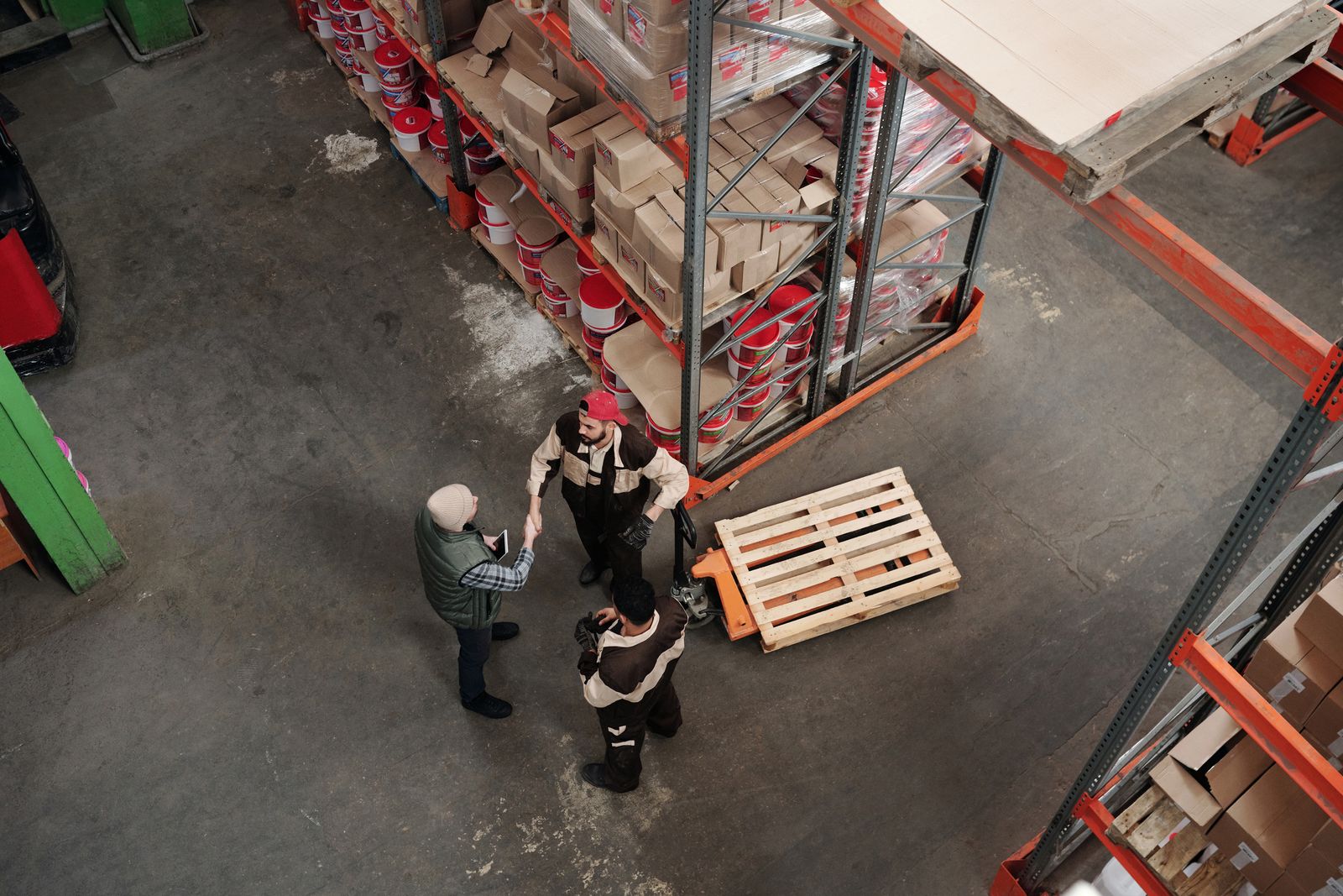 Three colleagues talk beside a pallet truck in a warehouse aisle, seen from above. Photo: Tiger Lily / Pexels.
