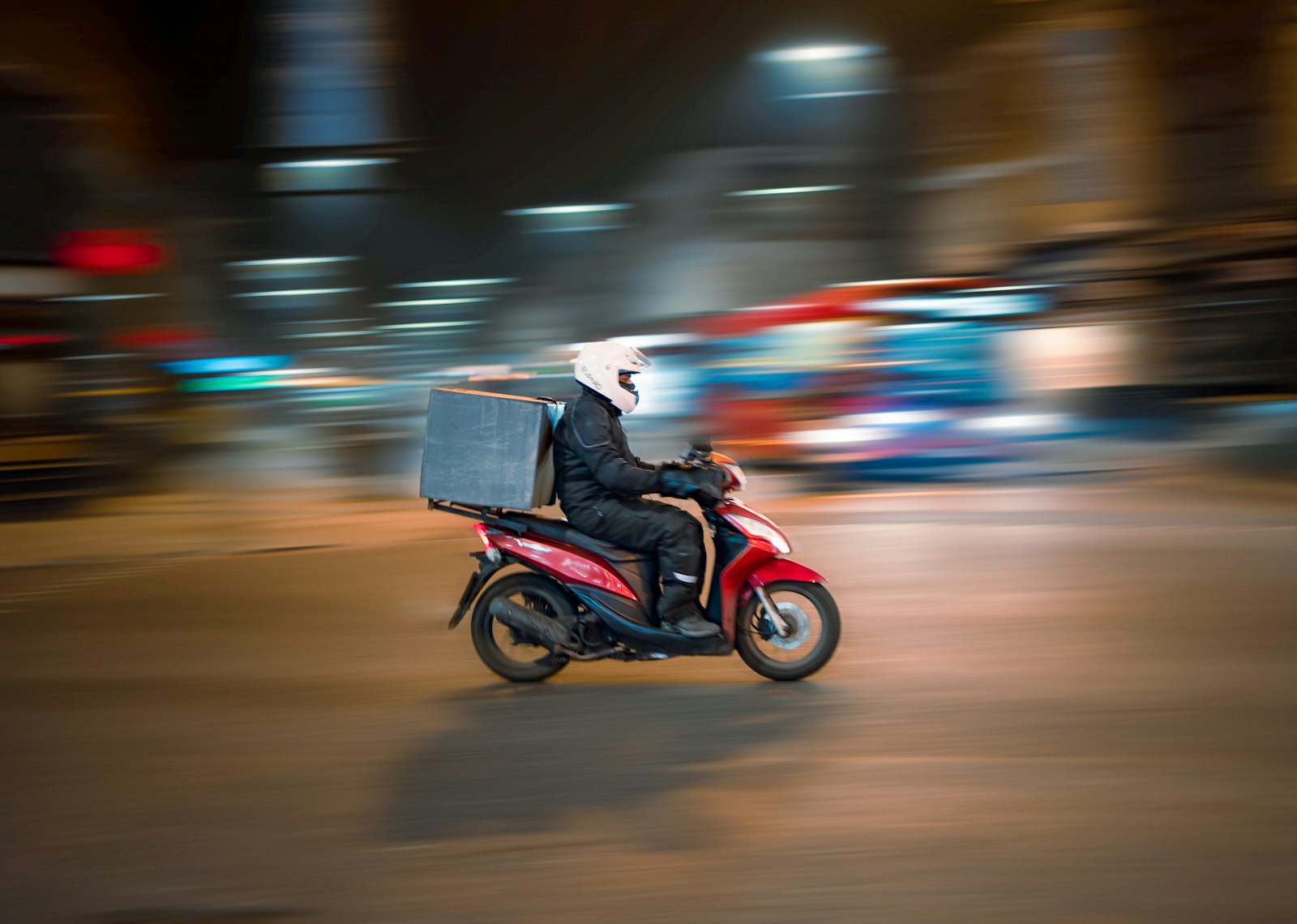 A delivery rider on a red scooter at night, a square cargo box on the rack, city lights streaking past. Photo: Rowan Freeman / Unsplash.