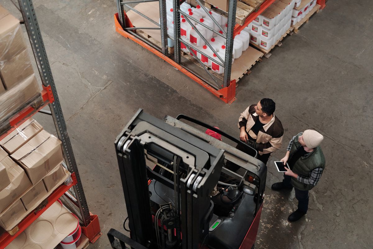 Two warehouse workers beside a reach truck look up at a loaded rack, one holding a tablet, seen from above. Photo: Tiger Lily / Pexels.