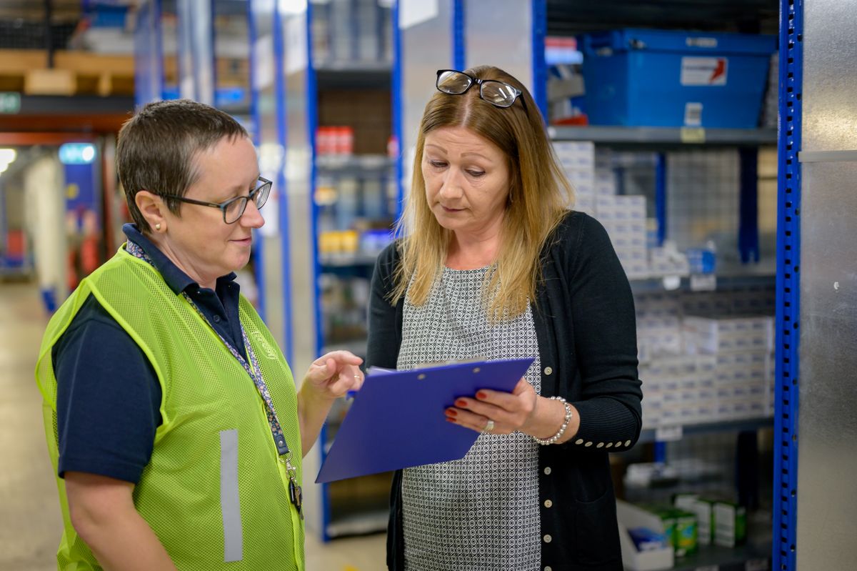 A supervisor in a hi-vis vest and a colleague go through a sheet on a clipboard between warehouse shelves. Photo: Centre for Ageing Better / Pexels.