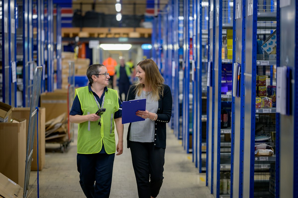 A supervisor with a clipboard walking a stockroom aisle beside a picker in a hi-vis vest holding a barcode scanner.